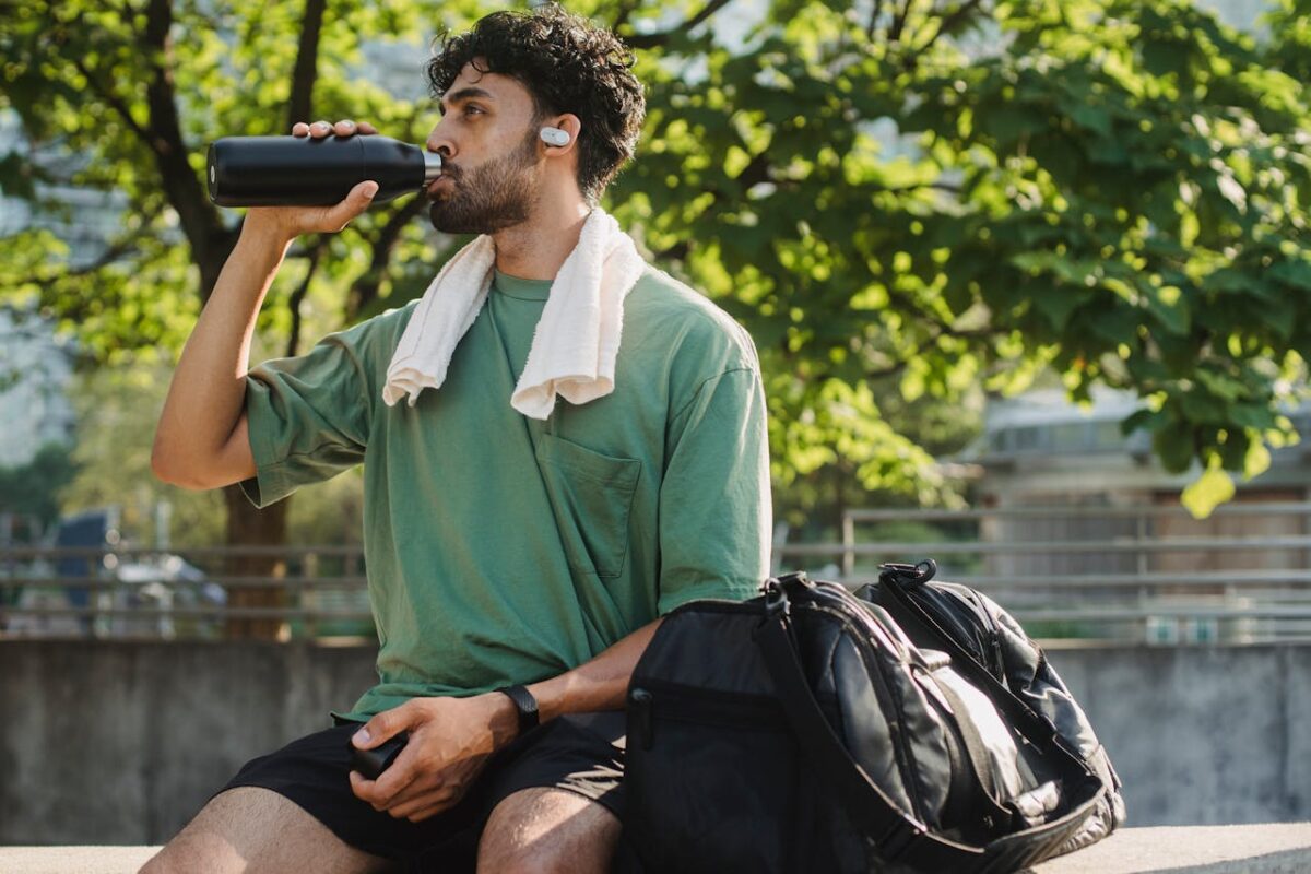 Man with green shirt and curly hair drinks water outdoors post-exercise.