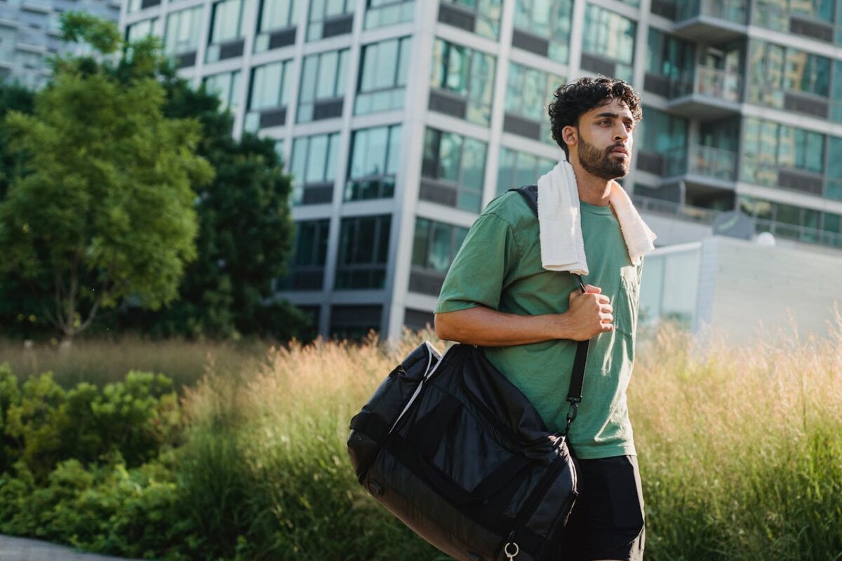 Young man with duffle bag and towel walking in urban park during summer.