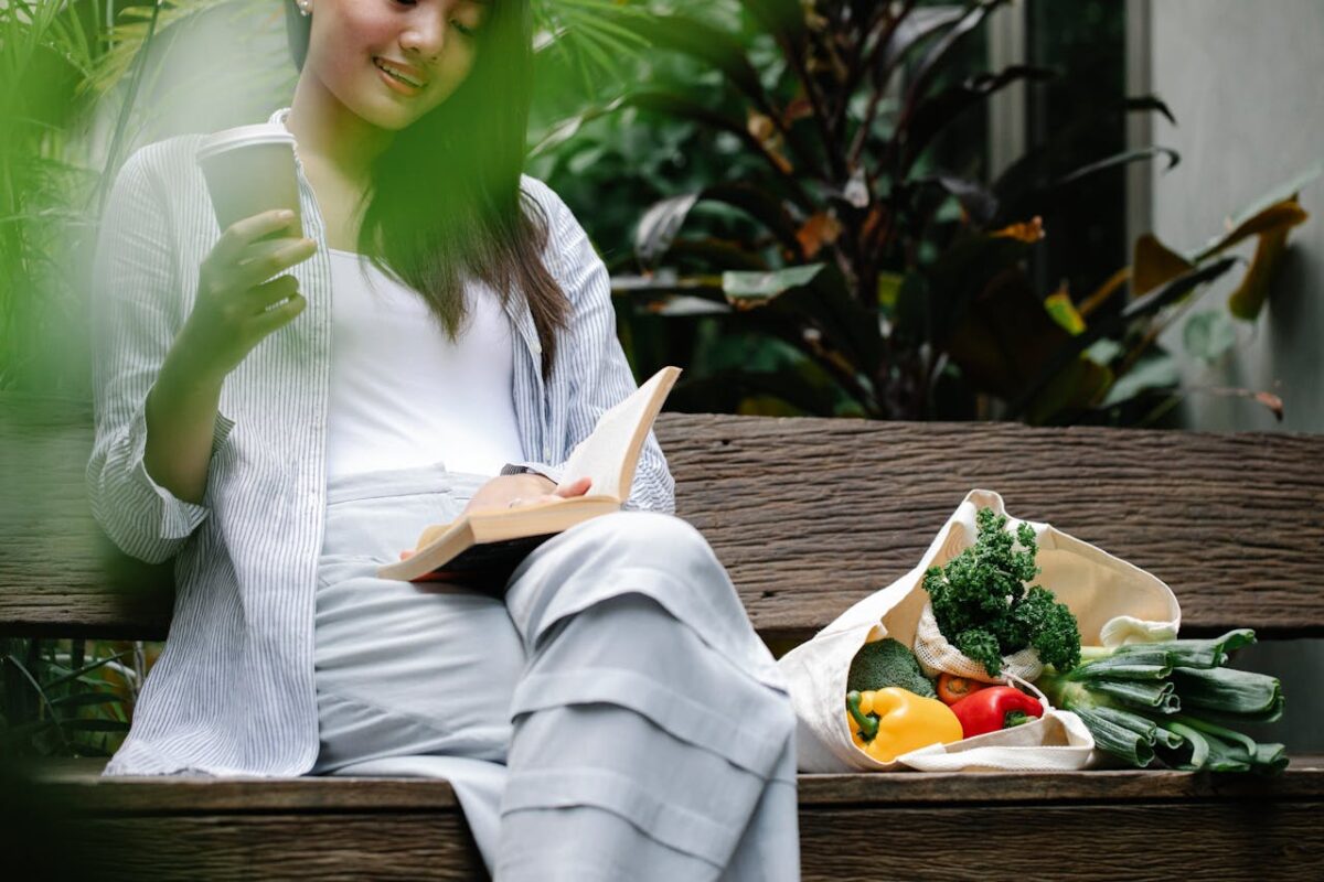 Crop smiling Asian female wearing casual outfit reading book and drinking takeaway coffee while sitting on bench with ripe vegetables in verdant garden